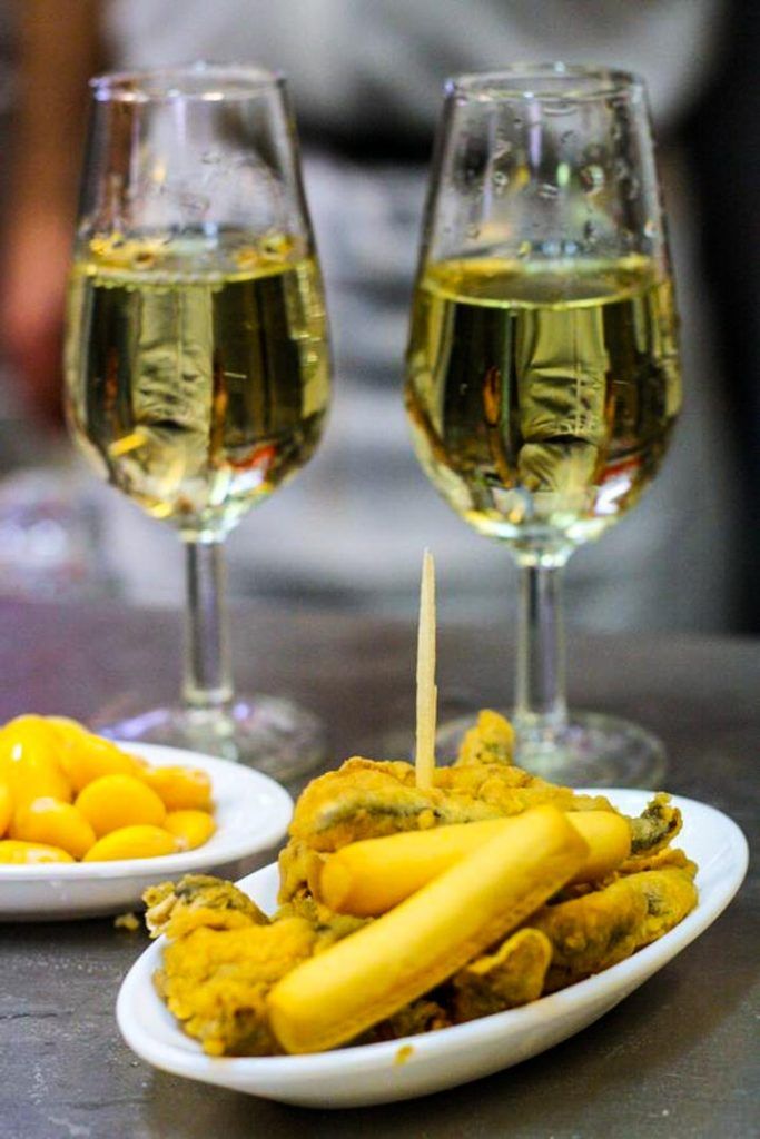 Dos copas de vino blanco con un plato de tapas en un bar, plan gastronómico para completar qué ver en Frigiliana en un día.