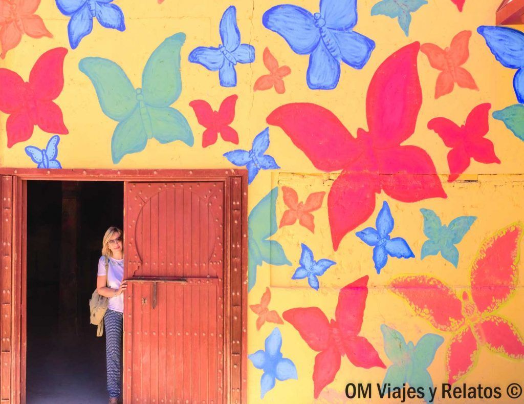 Foto mía asomándome por una puerta roja junto a un mural de mariposas de colores, una parada fotogénica en la ruta Ouarzazate desde Marrakech.