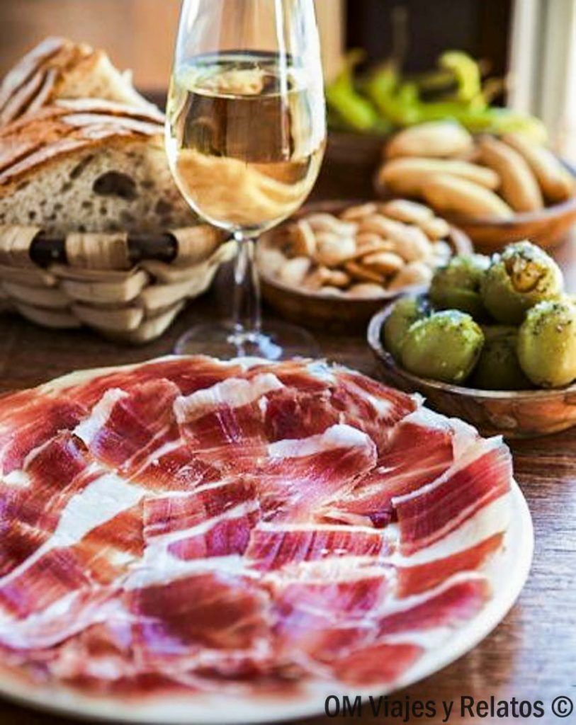 Plato de jamón ibérico con copa de vino, pan y aceitunas, experiencia gastronómica en pueblos más bonitos de Cáceres.