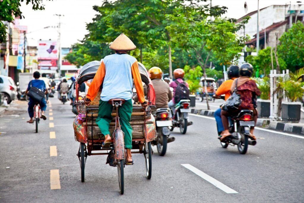 Ciclista conduciendo un becak tradicional por las calles de Yogyakarta, compartiendo espacio con motos y el tráfico local, una experiencia auténtica entre las mejores cosas que ver en Yogyakarta.