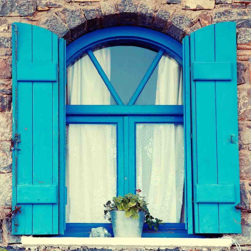 Ventana azul con contraventanas y maceta en una casa tradicional de Creta, un detalle con encanto entre las islas griegas mas bonitas.