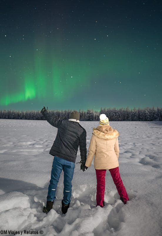 Foto mía y de JC de espaldas en la nieve señalando la aurora boreal en el cielo de, ideal para ver auroras boreales en Rovaniemi.