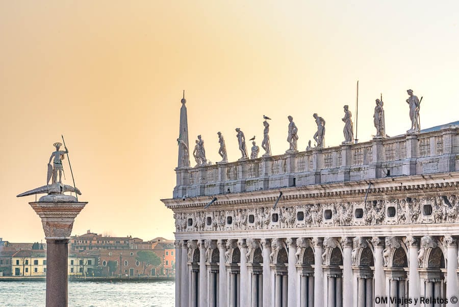 Detalle arquitectónico de las esculturas sobre la Biblioteca Marciana y la columna de San Teodoro al atardecer, una joya que visitar en la Plaza de San Marcos en Venecia.