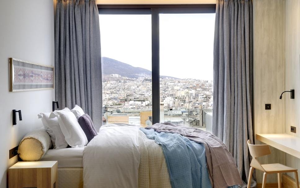 Habitación de hotel luminosa con ventanal y vistas a la ciudad, perfecta para imaginar donde alojarse en Atenas con buenas panorámicas.