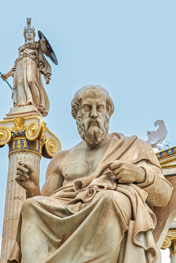 Estatua clásica frente a la Academia de Atenas con Atenea al fondo, una zona céntrica a considerar si buscas donde alojarse en Atenas.