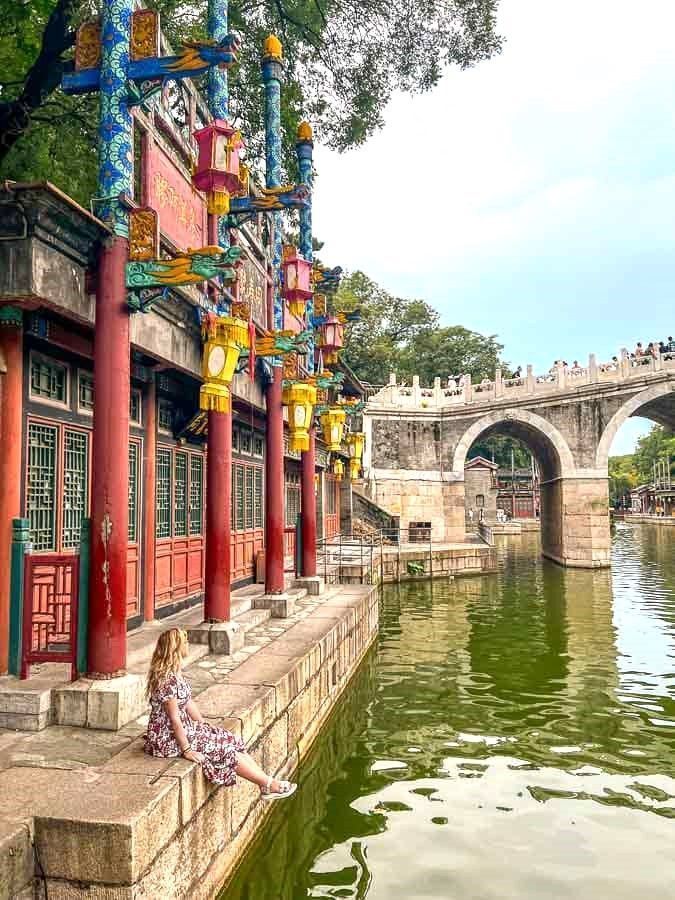 Foto mía sentada junto al canal del Palacio de Verano, con faroles y un puente de piedra al fondo, en la ruta de que ver en Pekín en 4 dias.