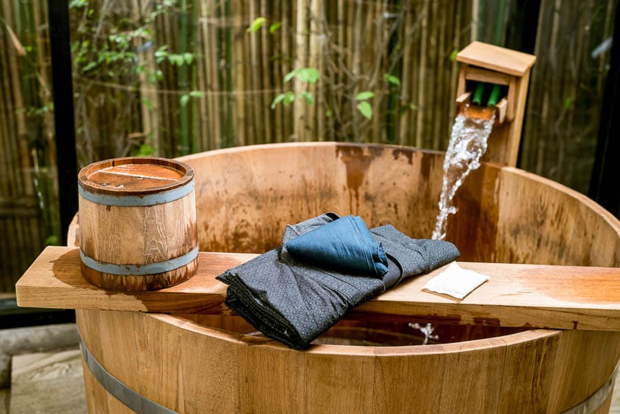 Bañera ofuro de madera con agua caliente cayendo y yukata doblada, ritual de relax en aguas termales en Japón.