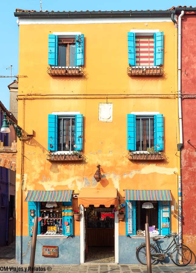 Fachada amarilla de Burano con contraventanas azules, tienda local y bicicleta, un clásico de que ver en Burano.