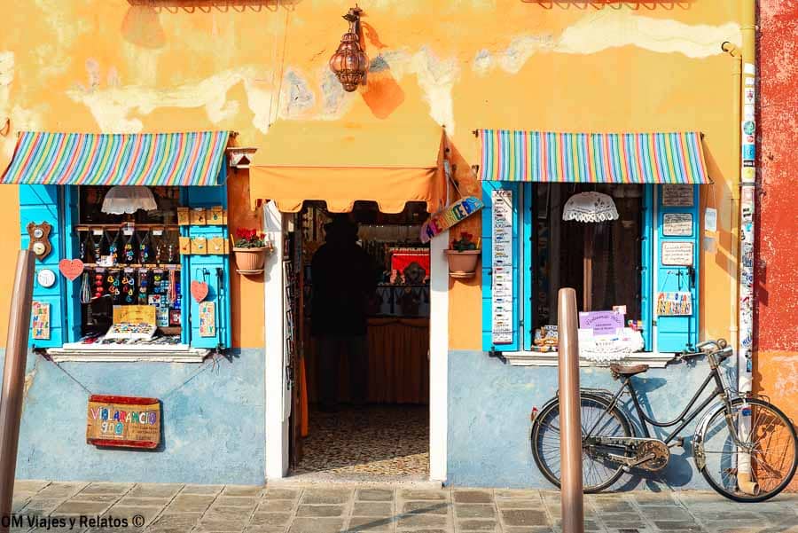 Fachada de tienda típica en Burano con ventanas azules, toldos de colores y bicicleta, un imprescindible en que ver en Burano.