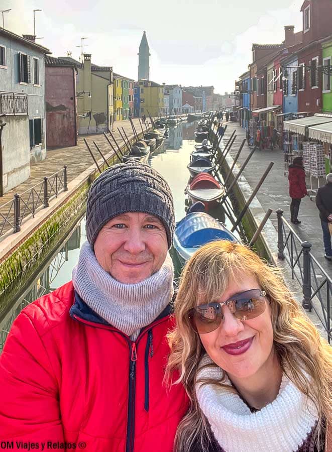 Selfie de pareja de OM Viajes y Relatos en un puente con el canal de Burano y casas de colores al fondo, una foto top de que ver en Burano.