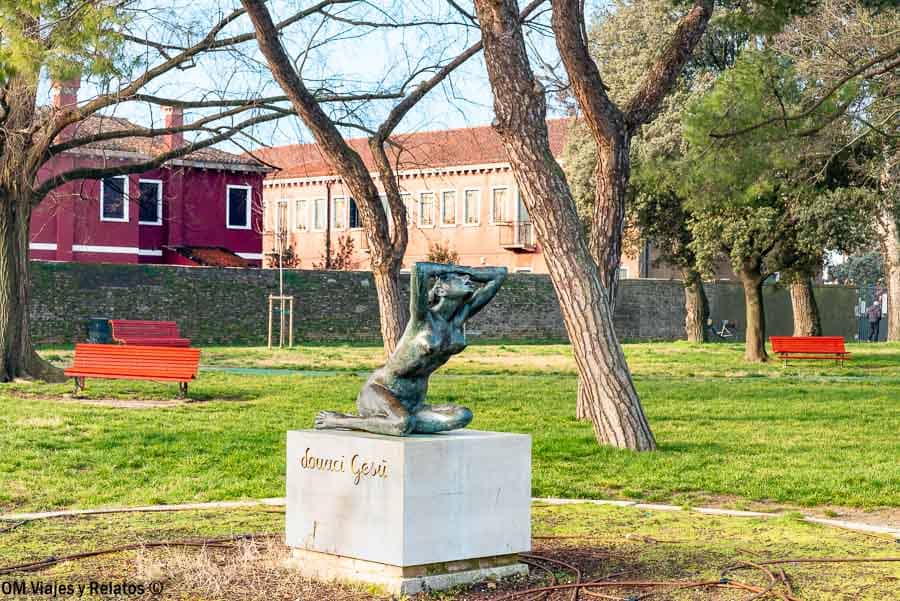 Escultura “Attesa di pace” en un parque de Burano entre árboles y bancos rojos, un rincón diferente de que ver en Burano.
