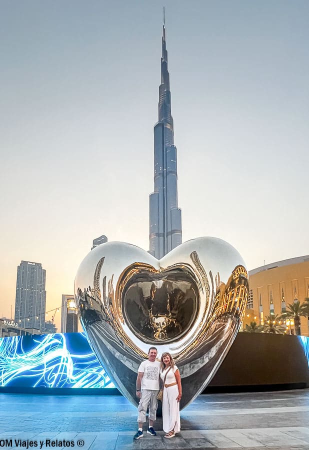 Subir al Burj Khalifa: Yo y JC posando frente a una escultura de corazón plateado con el Burj Khalifa al fondo al atardecer en Dubái.