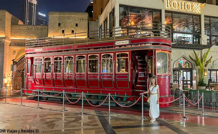 Una servidora junto a un tranvía turístico rojo en una zona comercial de Dubái de noche, una experiencia urbana para incluir en los consejos para viajar a Dubái.