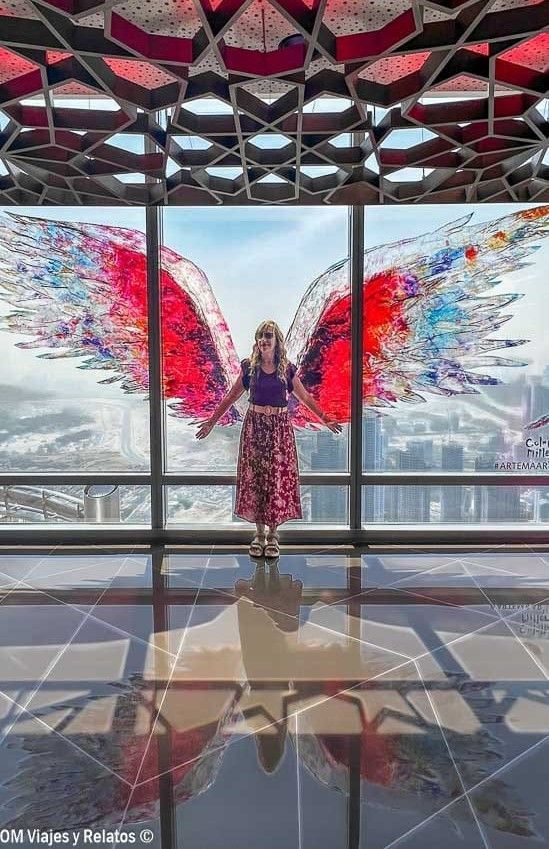 Foto mía posando frente a un mural de alas rojas en un mirador acristalado de Dubái, durante la experiencia de subir al Burj Khalifa.