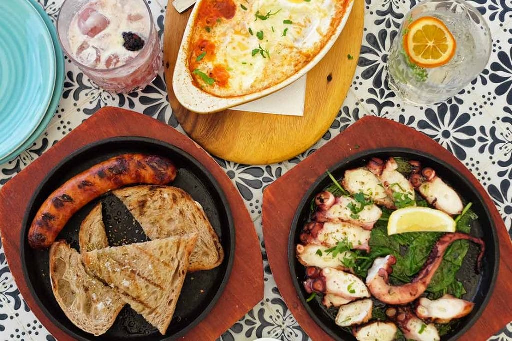 Mesa con platos típicos griegos, pulpo, salchicha y queso al horno, una experiencia gastronómica que disfrutar en una ruta sobre que ver en Atenas en 2 dias.