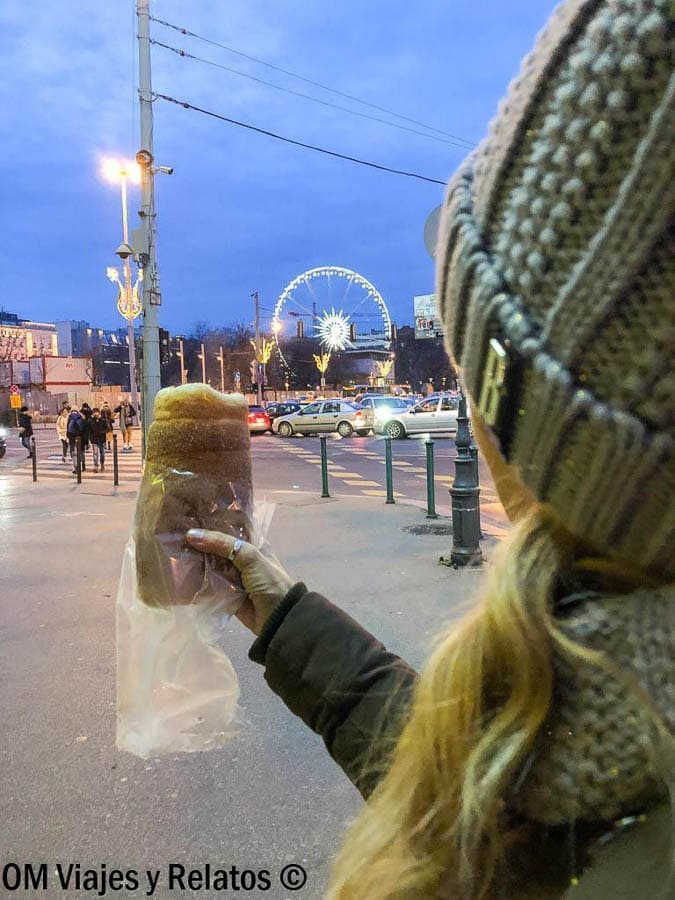 Una servidora con kürtőskalács frente a la noria iluminada de Budapest – Dulce típico en Budapest en Navidad.