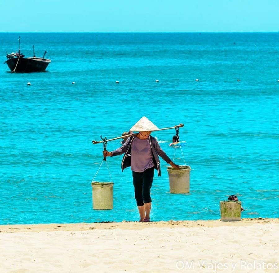 Mujer vietnamita con sombrero cónico llevando cubos en la playa, escena de vida local en Vietnam.