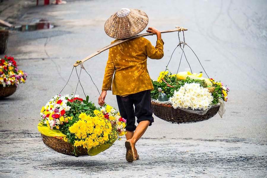 Vendedora ambulante de flores en Vietnam con sombrero cónico y cestas tradicionales en la calle.