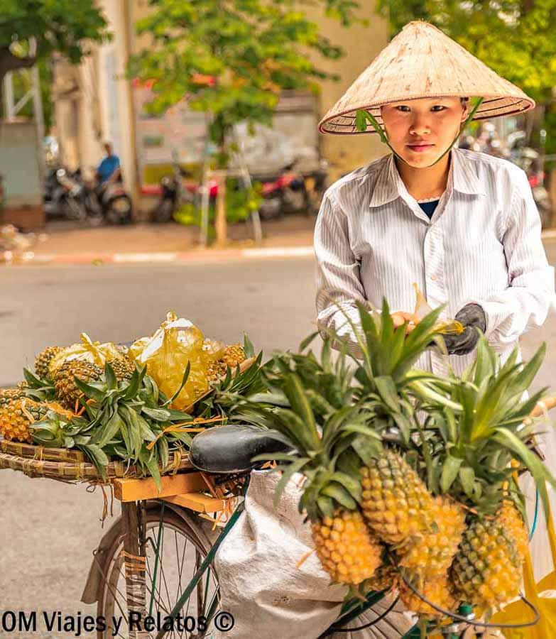 Vendedor ambulante de piñas en bicicleta con sombrero cónico, escena callejera imprescindible en Vietnam.