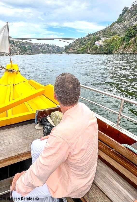 JC sentado en un barco navegando por el río Duero con puente al fondo y laderas verdes, una experiencia diferente que ver en Oporto en 2 días.