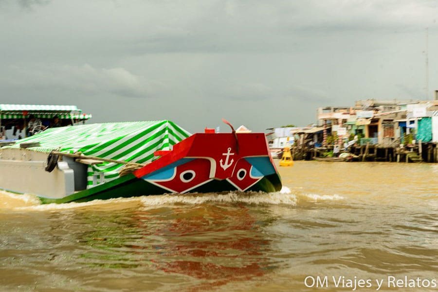 Delta del Mekong Vietnam: barco tradicional navegando por el río frente a casas flotantes y mercado local.
