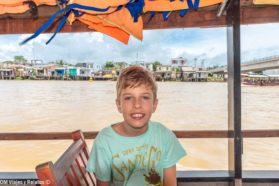 Delta del Mekong Vietnam: Raúl en un barco tradicional con el río y casas junto a la orilla al fondo.