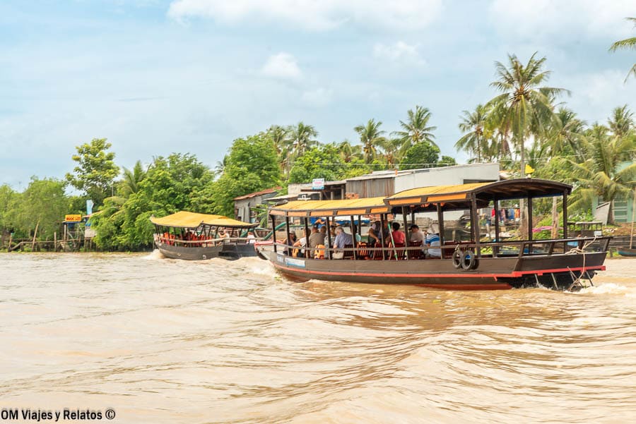 Delta del Mekong Vietnam: barco turístico navegando por el río marrón con palmeras y casas en la orilla.