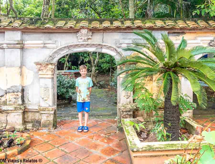 Nuestro hijo Raúl posa bajo un arco de piedra en un patio con baldosas rojas, muro antiguo y palmera en macetero, rodeado de vegetación tropical.