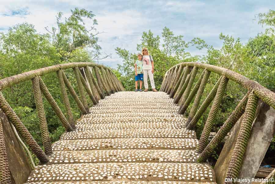 Yo y mi hijo Raúl posan al final de un puente curvo de piedra con barandillas trenzadas, rodeado de vegetación tropical en el Delta del Mekong, Vietnam