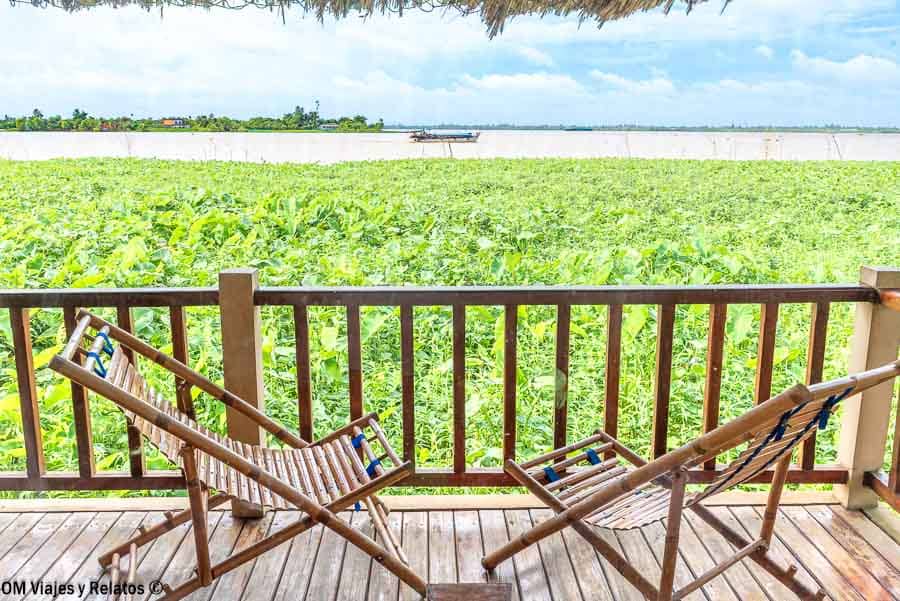 Terraza del Mekong Riverside Resort con dos sillas de madera frente al río Mekong cubierto de plantas acuáticas, con una barca al fondo, en el Delta del Mekong (Vietnam).