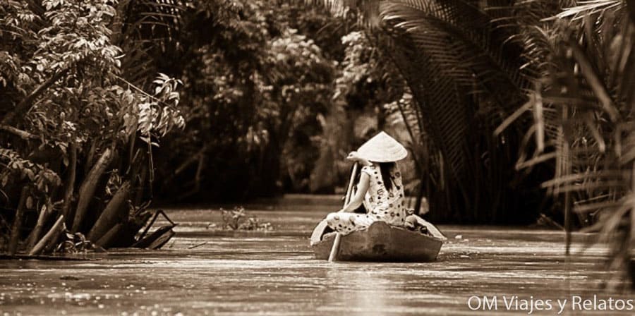 Delta del Mekong Vietnam: mujer con sombrero cónico remando una pequeña barca por un canal rodeado de vegetación.