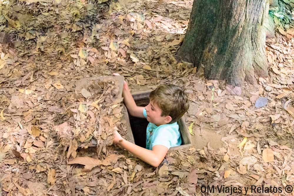 Nuestro hijo saliendo por una trampilla camuflada entre hojas secas en los túneles de Củ Chi, Vietnam.
