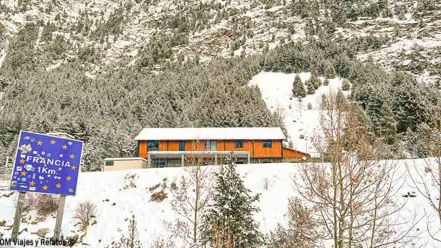 Cartel de “Francia 1 km” junto a un paisaje nevado de montaña y bosque, foto tomada a un kilómetro de la frontera rumbo a los pueblos más bonitos del Pirineo Francés.