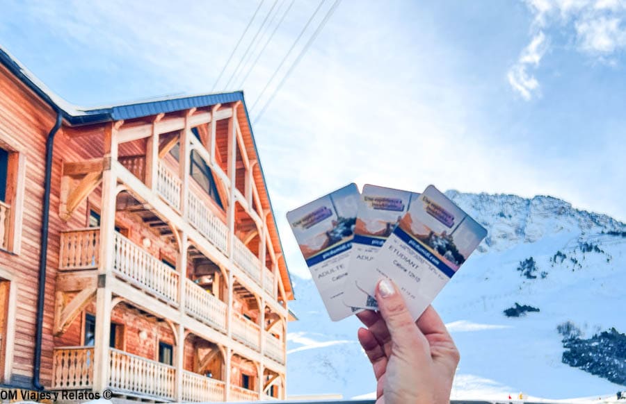 Entradas para subir al Pic du Midi sostenidas por mi, con edificio de montaña y paisaje nevado al fondo, una excursión cerca de los pueblos más bonitos del Pirineo Francés.