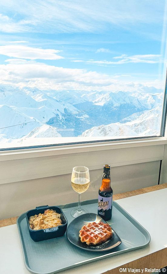Snack en el Pic du Midi con gofre y bebida en una bandeja frente a un ventanal con vistas a las montañas nevadas, una experiencia cerca de los pueblos más bonitos del Pirineo Francés.