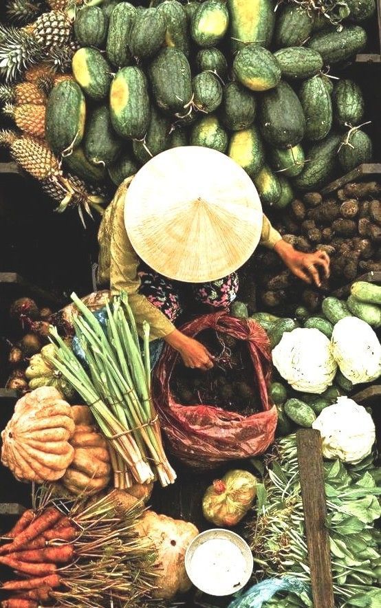 Delta del Mekong Vietnam: mercado local con vendedora con sombrero cónico rodeada de frutas y verduras tropicales.