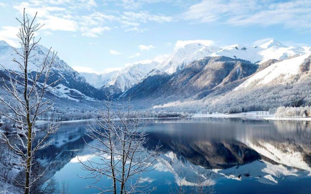 Vista invernal del Lago de Génos-Loudenvielle con montañas nevadas y árboles sin hojas frente al agua, cerca de los pueblos más bonitos del Pirineo Francés.