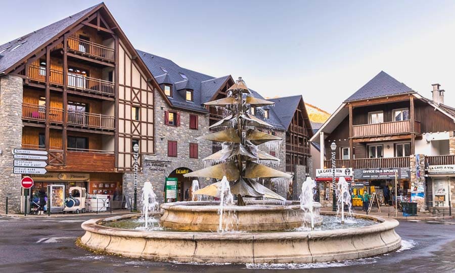 Plaza con fuente escultórica y edificios de madera y piedra en Saint-Lary-Soulan, uno de los pueblos más bonitos del Pirineo Francés.