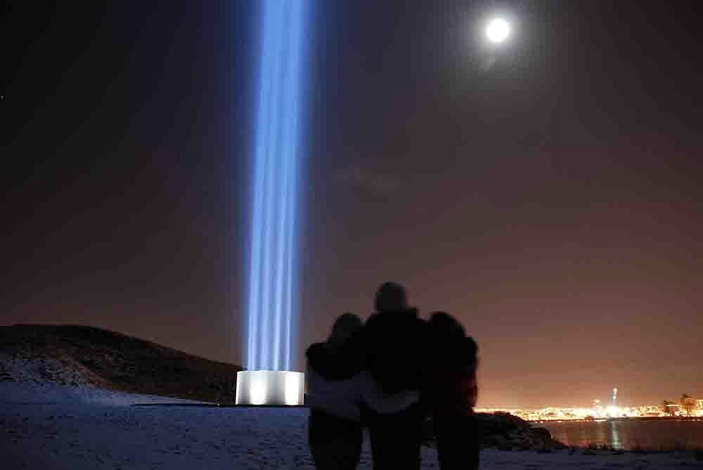 Pareja abrazada frente a la Imagine Peace Tower iluminando el cielo en Reikiavik, una escena increíble de Islandia en Octubre.