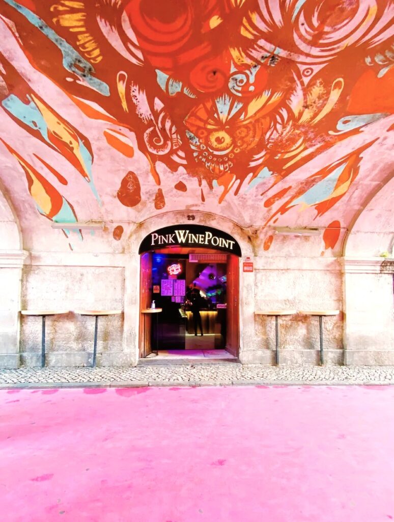Entrada colorida de Pink Wine Point en Lisboa con techo de arte urbano y suelo rosa, una zona llamativa a valorar al decidir donde alojarse en Lisboa.