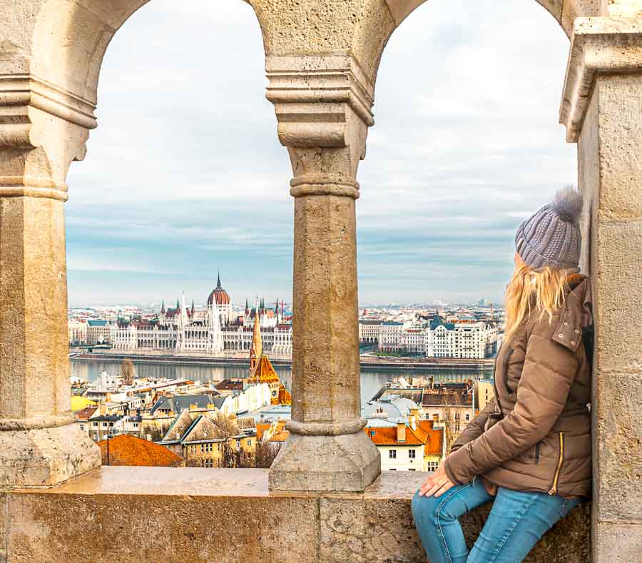 Olga en un mirador de Budapest con el Parlamento al fondo entre columnas de piedra, una imagen perfecta para inspirar un viaje a Budapest por libre.