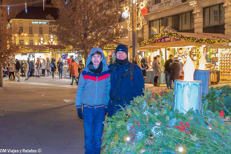 JC y nuestro hijo en un mercado navideño de Budapest con puestos iluminados al fondo