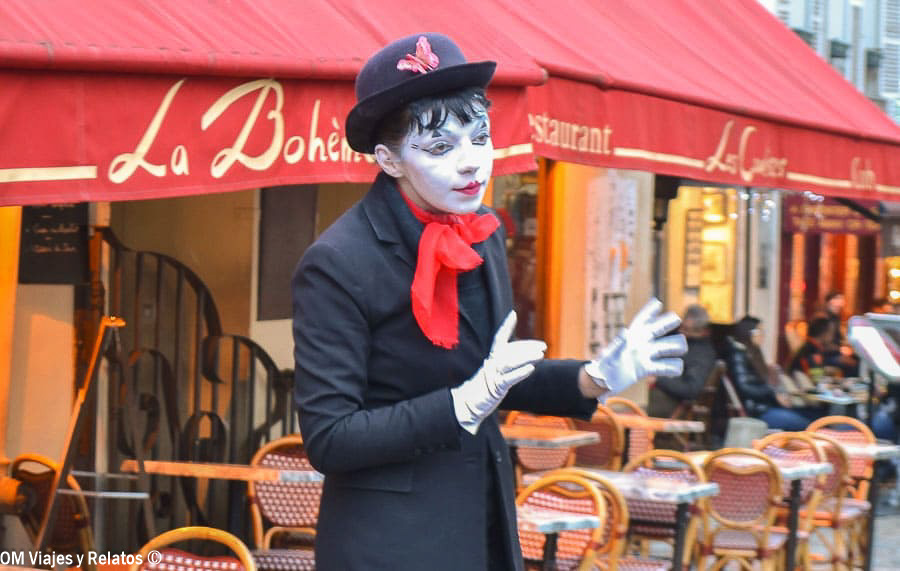 Artista callejero caracterizado como mimo, con sombrero negro y pañuelo rojo, frente al restaurante La Bohème en Montmartre, París