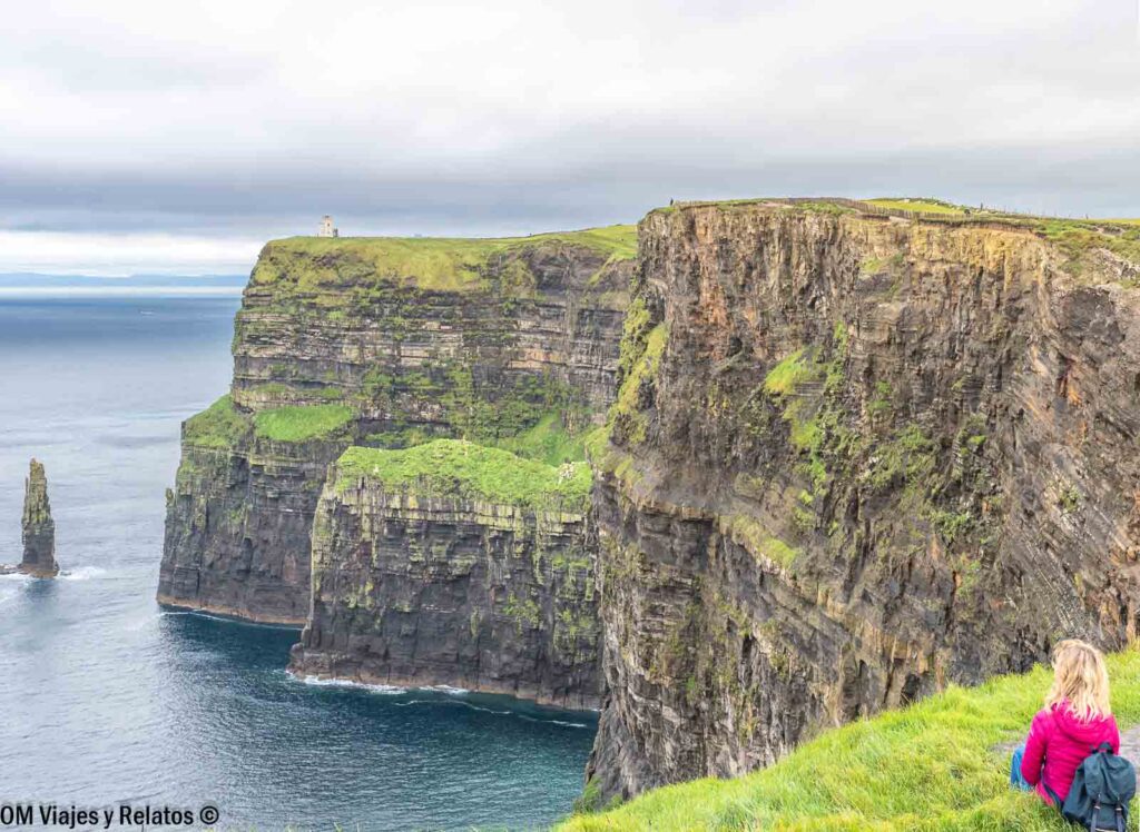 Una servidora sentada sobre una pradera verde contemplando las enormes paredes rocosas de los Acantilados de Moher y el océano Atlántico