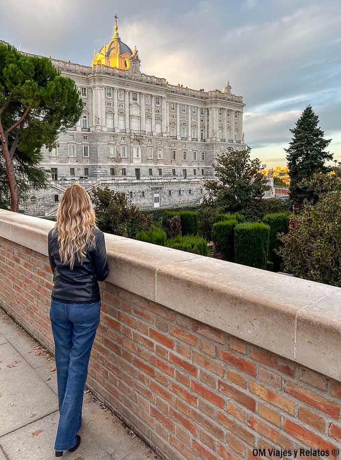 Servidora contemplando el Palacio Real de Madrid al atardecer, con su cúpula iluminada y los jardines en primer plano