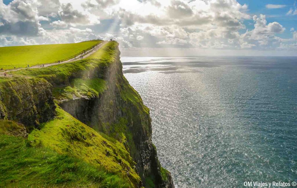 Vista de los Acantilados de Moher iluminados por el sol, con verdes praderas, un sendero junto a la cima y el océano Atlántico