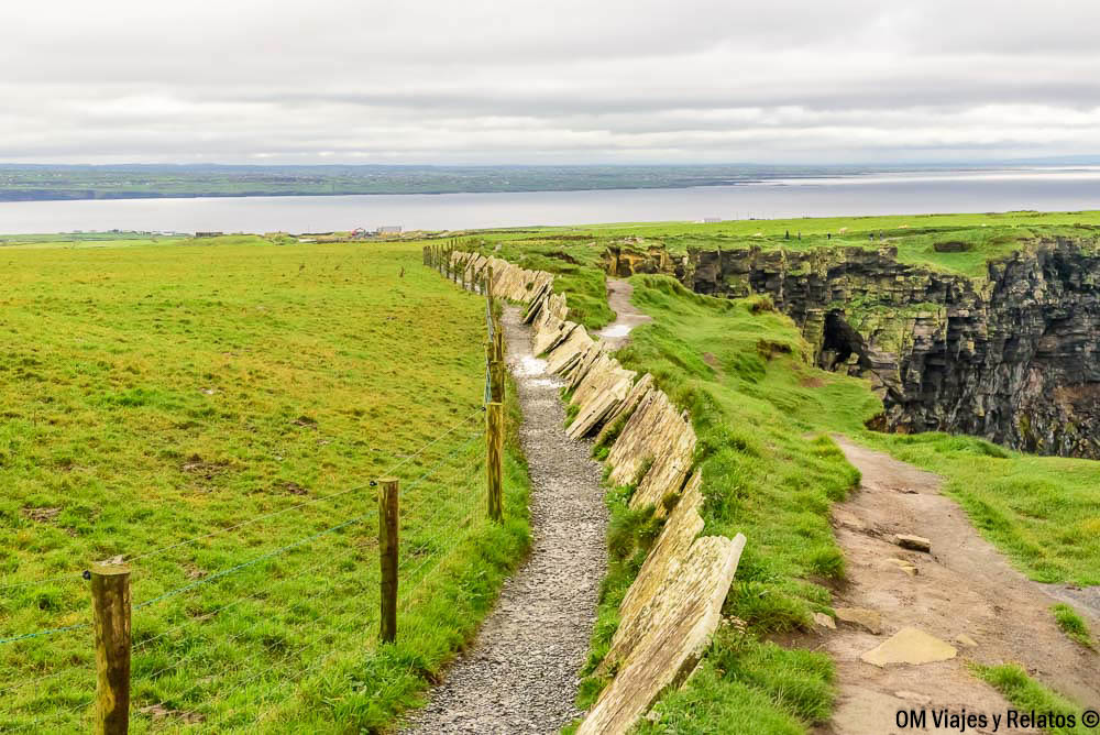 Sendero de tierra junto a los Acantilados de Moher, bordeado por una valla de piedra, praderas verdes y vistas al océano Atlántico